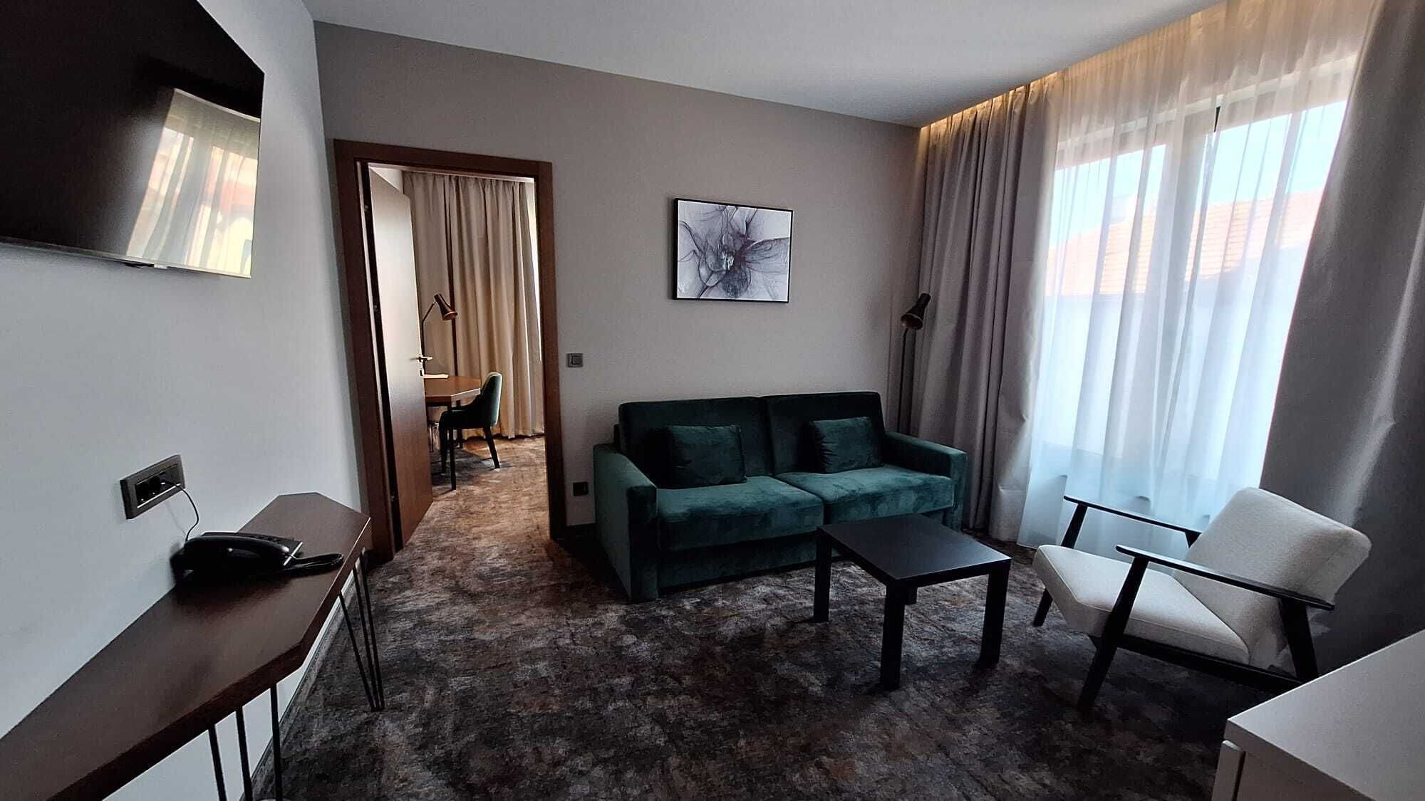 A modern hotel room with a green sofa, a small table, a chair, a flat-screen TV, and a large window with curtains.