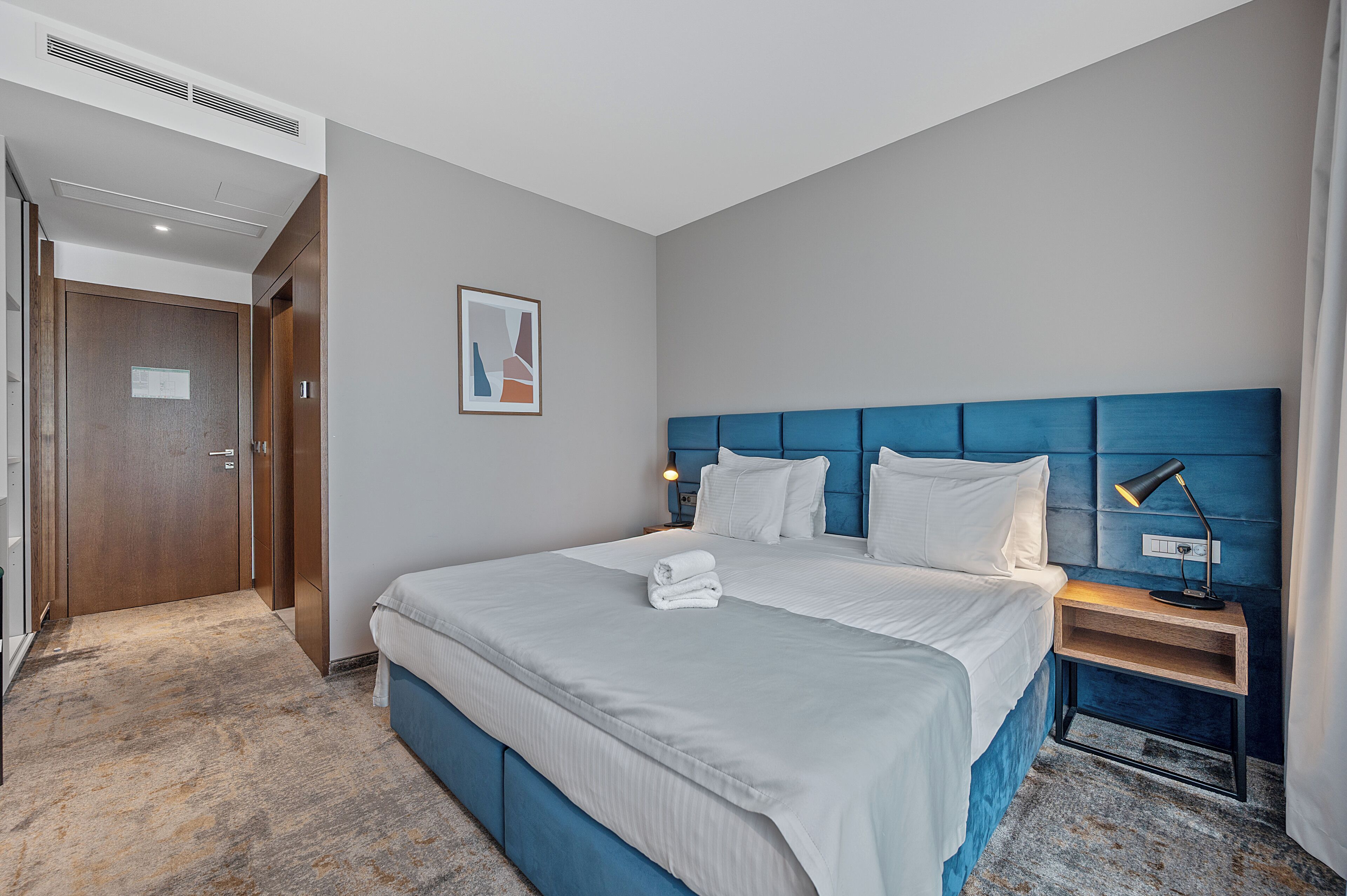 Quarto de hotel com uma cama grande, mesa de cabeceira com abajur, quadro na parede e porta à esquerda.