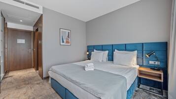 Quarto de hotel com uma cama grande, mesa de cabeceira com abajur, quadro na parede e porta à esquerda.