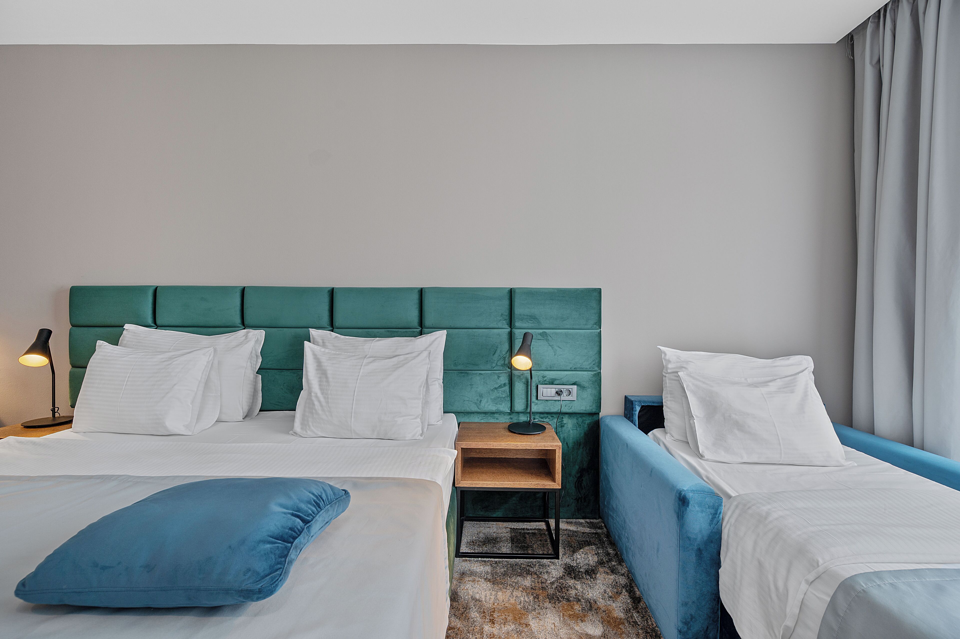 Habitación de hotel con dos camas, cabecera verde, ropa de cama blanca, almohada decorativa azul, mesita de noche con lámpara y ventana con cortinas.