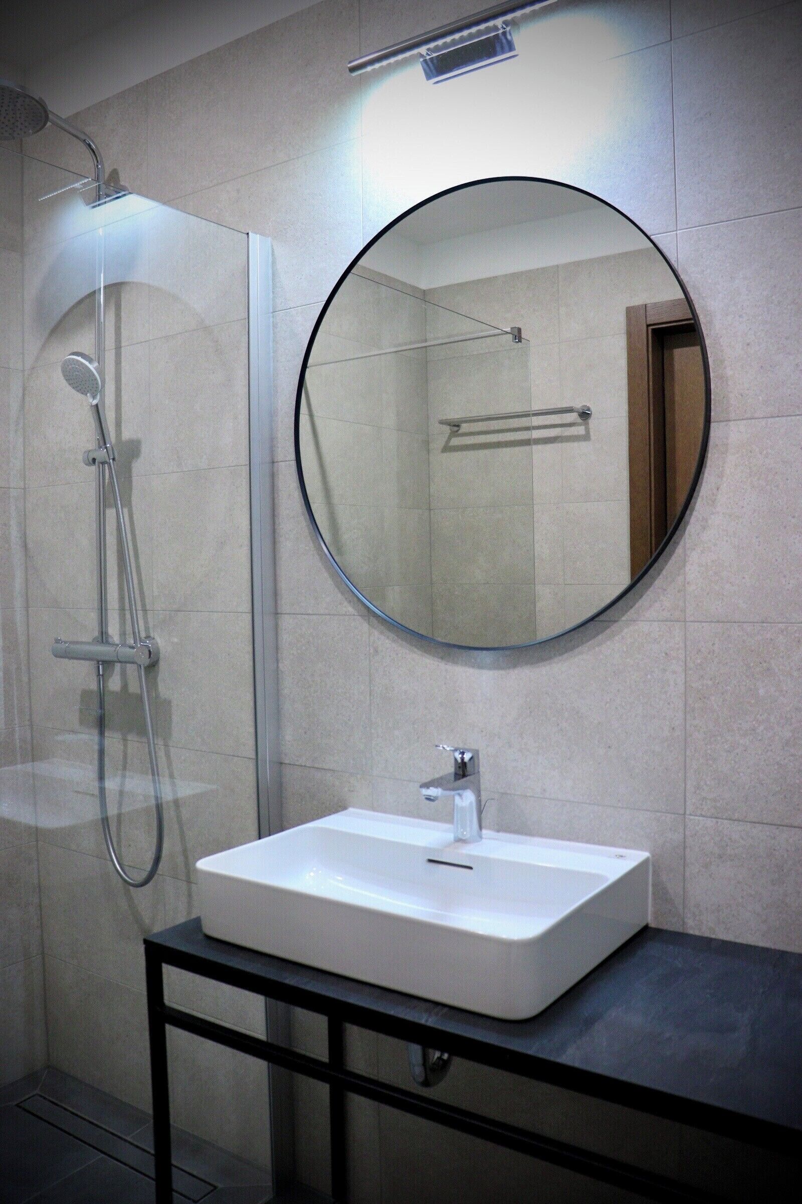 Un baño moderno con ducha de vidrio, espejo redondo y lavamanos blanco sobre encimera negra.