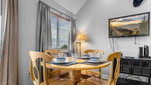 Een eethoek met een ronde houten tafel, zes houten stoelen, een televisie aan de muur en een raam met zonwering.