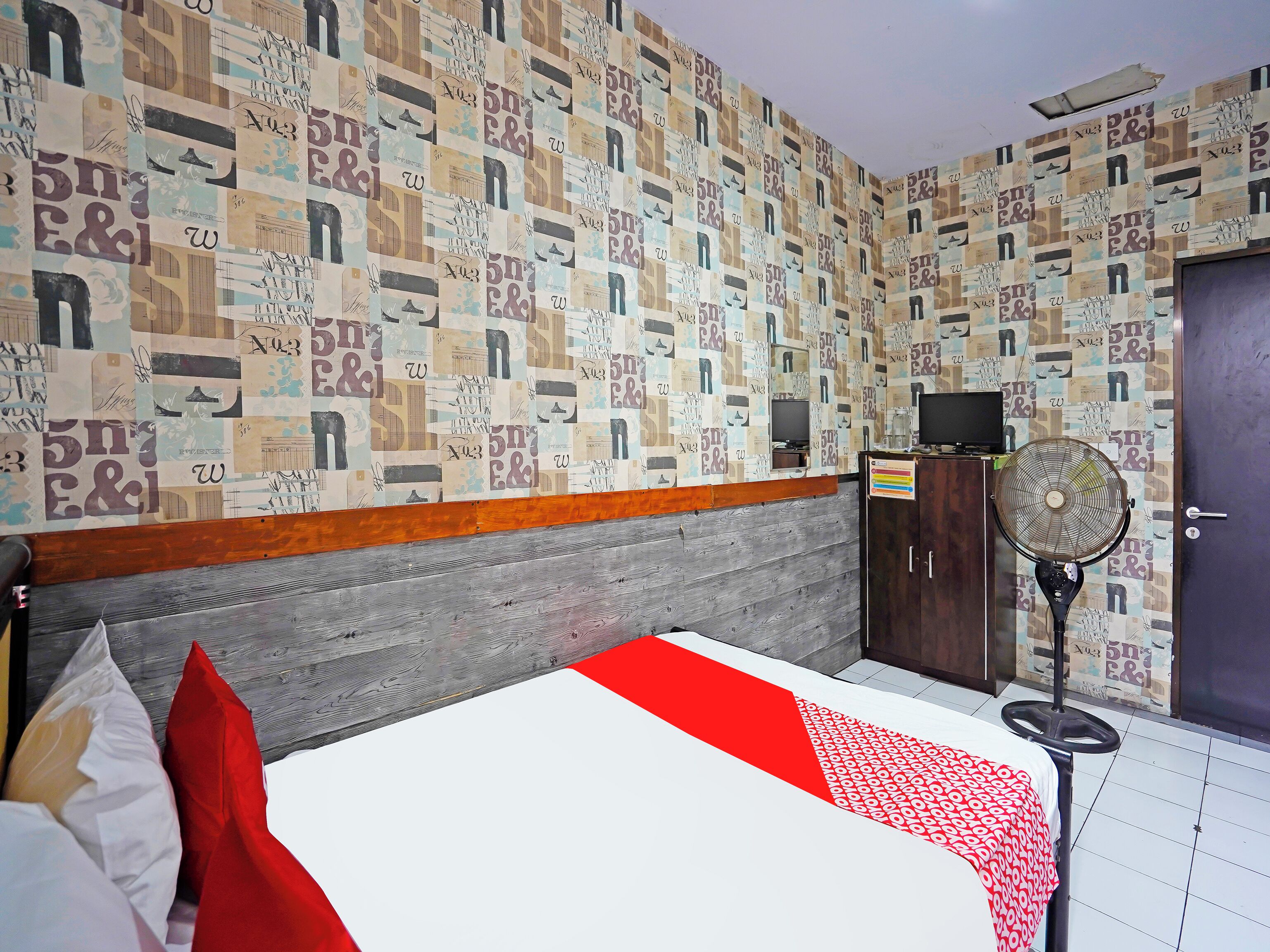 A hotel room with a patterned wall, a bed with a red and white bedspread, a wooden headboard, a wardrobe, a desk with a computer, and a standing fan.