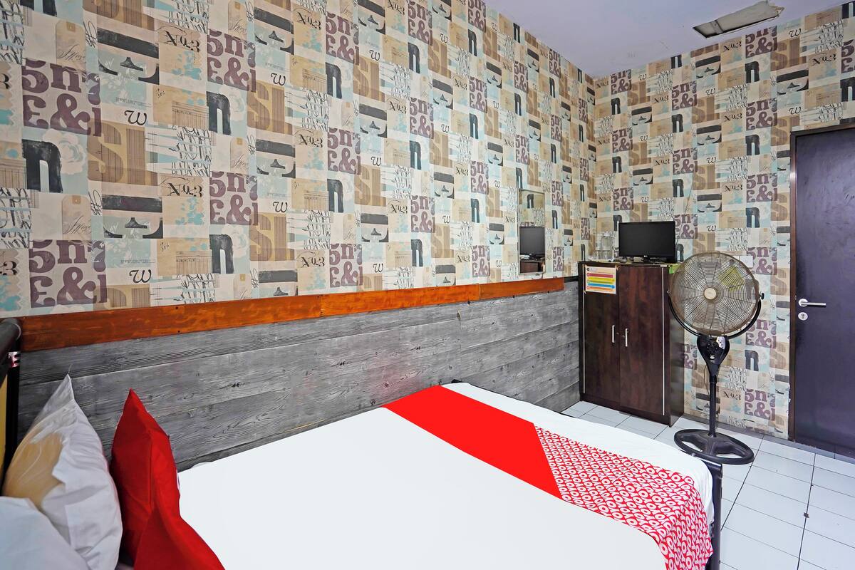 A hotel room with a patterned wall, a bed with a red and white bedspread, a wooden headboard, a wardrobe, a desk with a computer, and a standing fan.