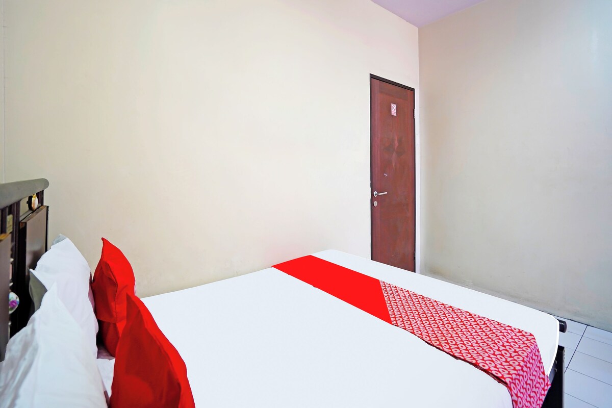 A hotel room with a bed, a wooden nightstand, a red door, and a white wall.