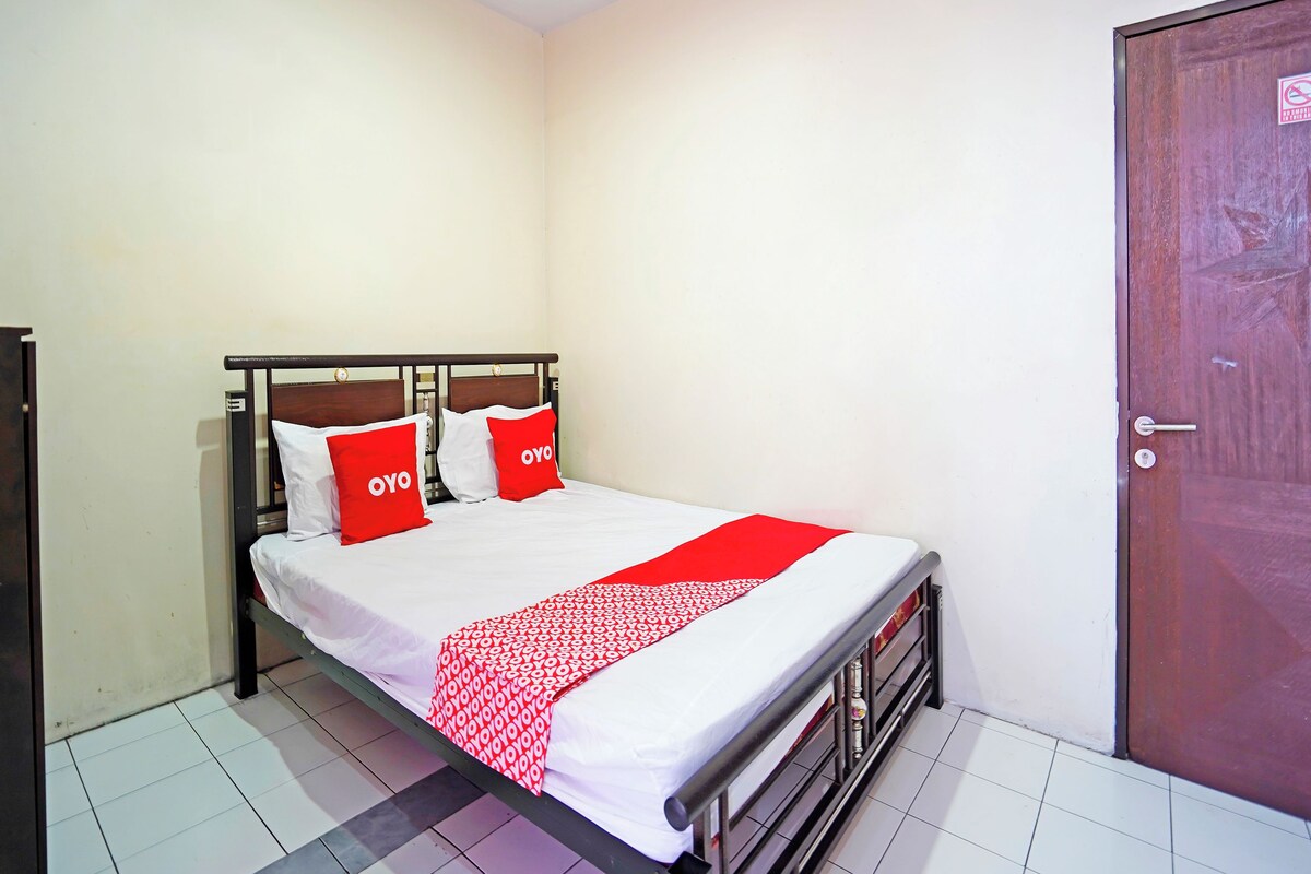 A single bed with a white and red bedspread, two red pillows with "OYO" text, a wooden door, and a tiled floor.