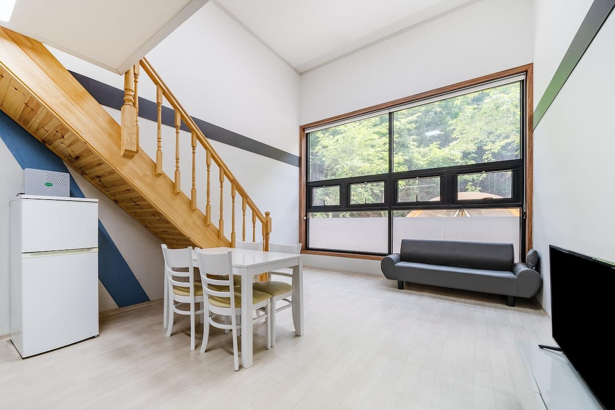 A modern living space with a wooden staircase, a dining area with white chairs, a sofa, and a large window offering a view of trees.