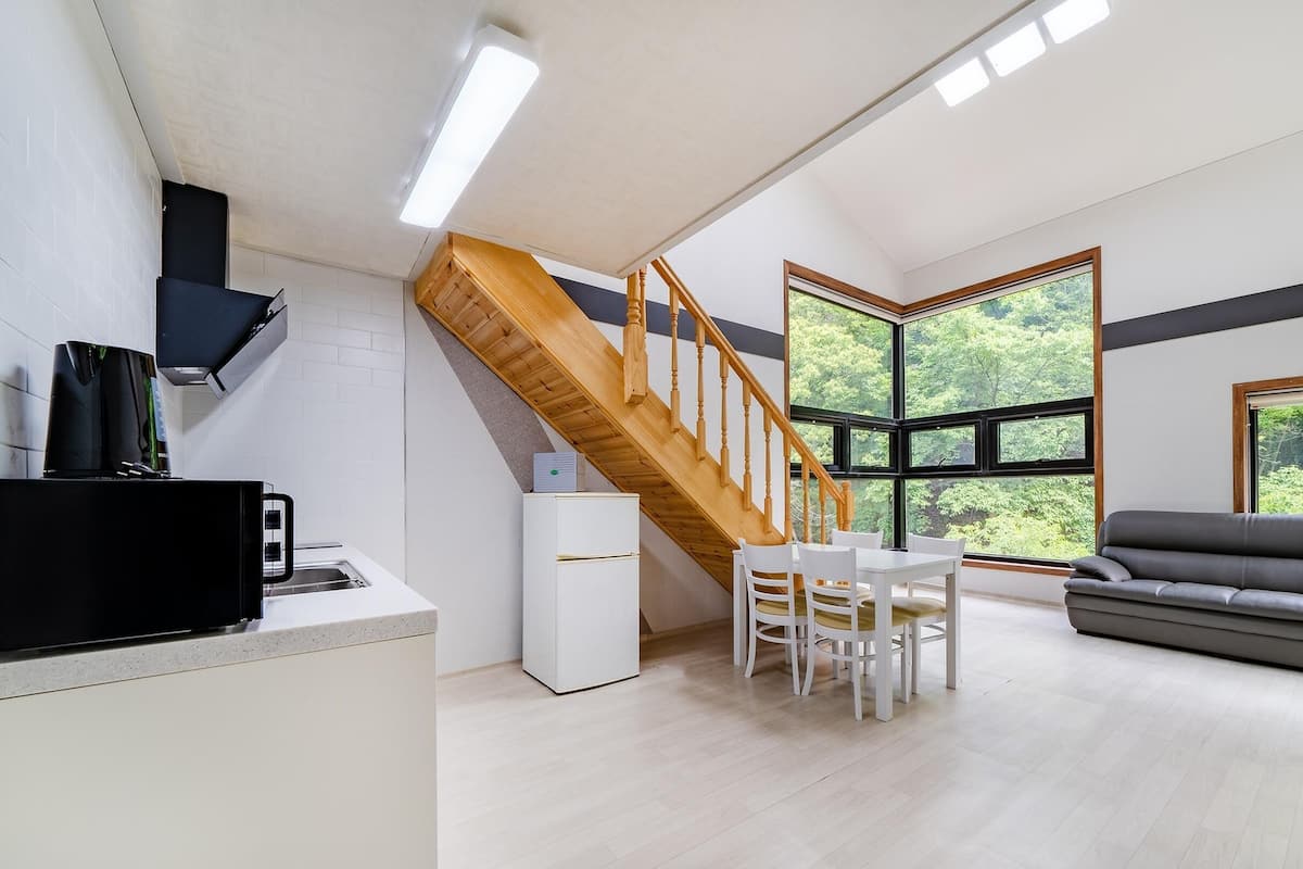 A modern kitchen with a built-in stove, a white refrigerator, and a dining area with a table and chairs. There is a staircase leading to a lofted area with a large window offering a view of greenery.