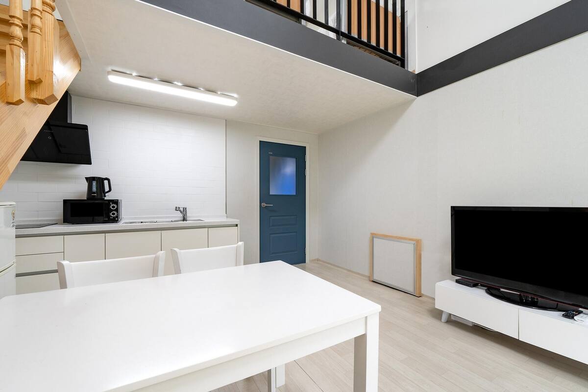 A modern kitchen with white cabinets, a black microwave, and a coffee maker. There's a blue door and a TV on a white stand.