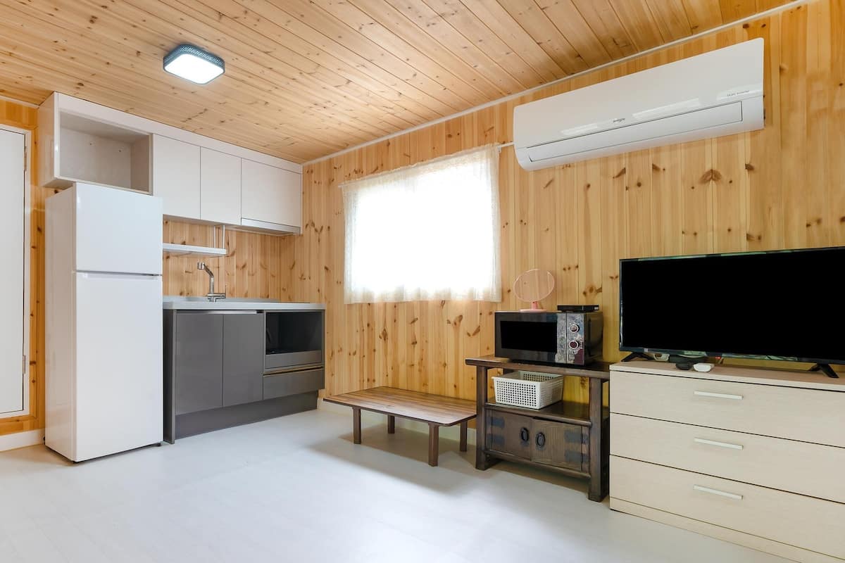 A compact kitchen with a microwave, stove, and sink, a small bench, and a TV on a wooden cabinet.