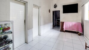 Une pièce spacieuse avec un sol carrelé de carreaux blancs, une porte fermée, un téléviseur fixé au mur et une petite table recouverte d’une nappe rose.