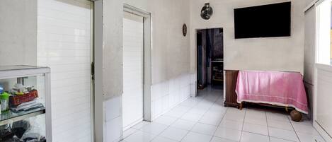 A spacious room with white tiled floors, a closed door, a wall-mounted TV, and a small table with a pink cloth.