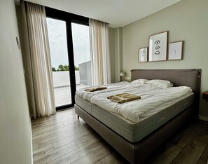 A modern bedroom with a large bed, a sliding glass door to the balcony, and a wall with framed artwork.