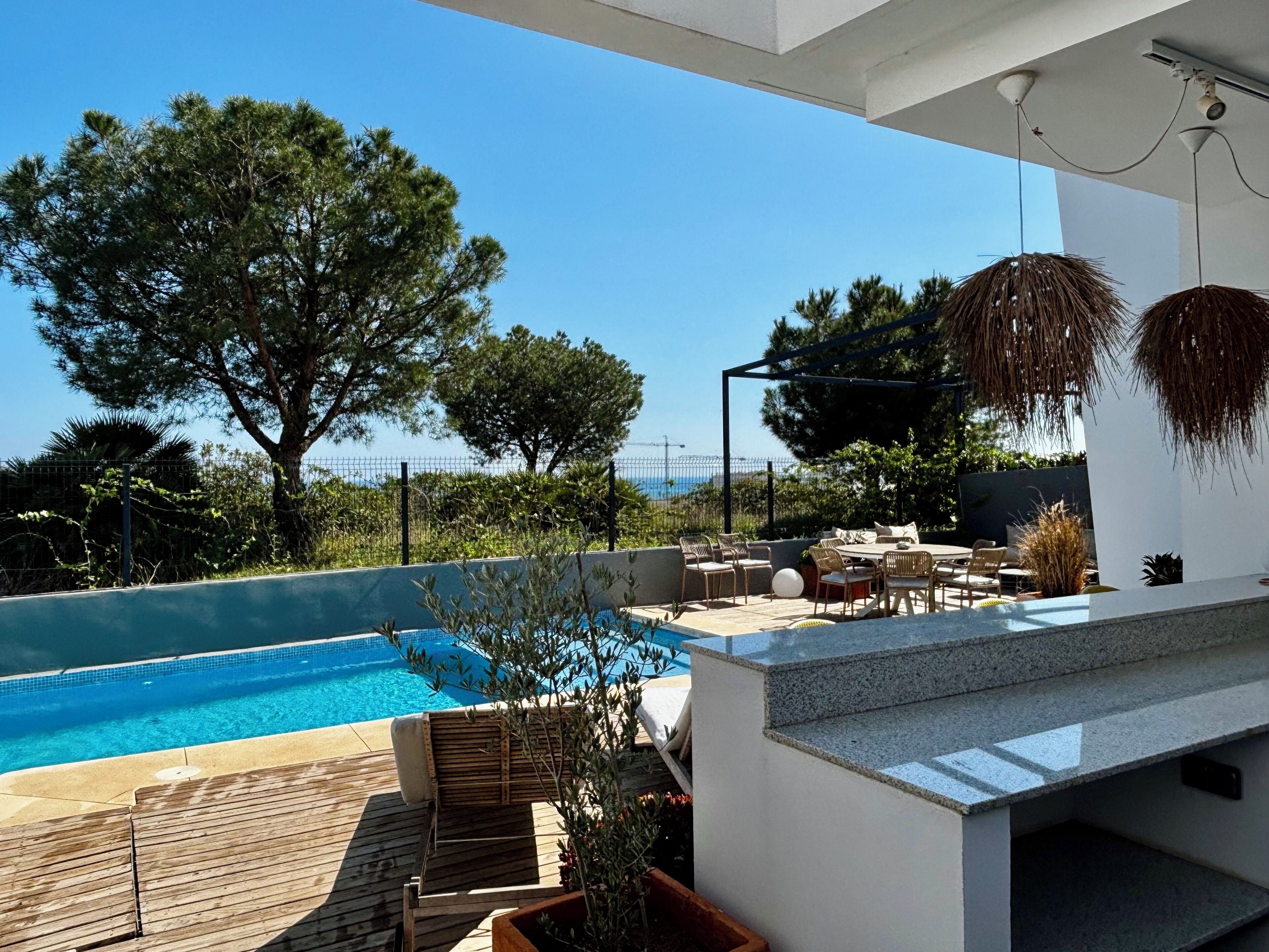 Zona junto a la piscina con vista al mar, mobiliario exterior y una pérgola.