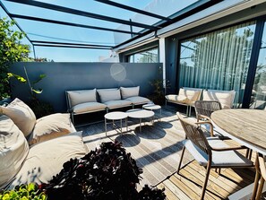 A modern outdoor patio with a glass enclosure, cushioned seating, and a wooden table.