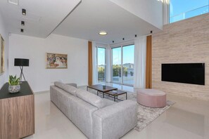 Ein modernes Wohnzimmer mit grauem Sofa, Couchtisch, pinkem Hocker, flachbildschirm-Fernseher an der Wand und einem großen Fenster mit Vorhängen.