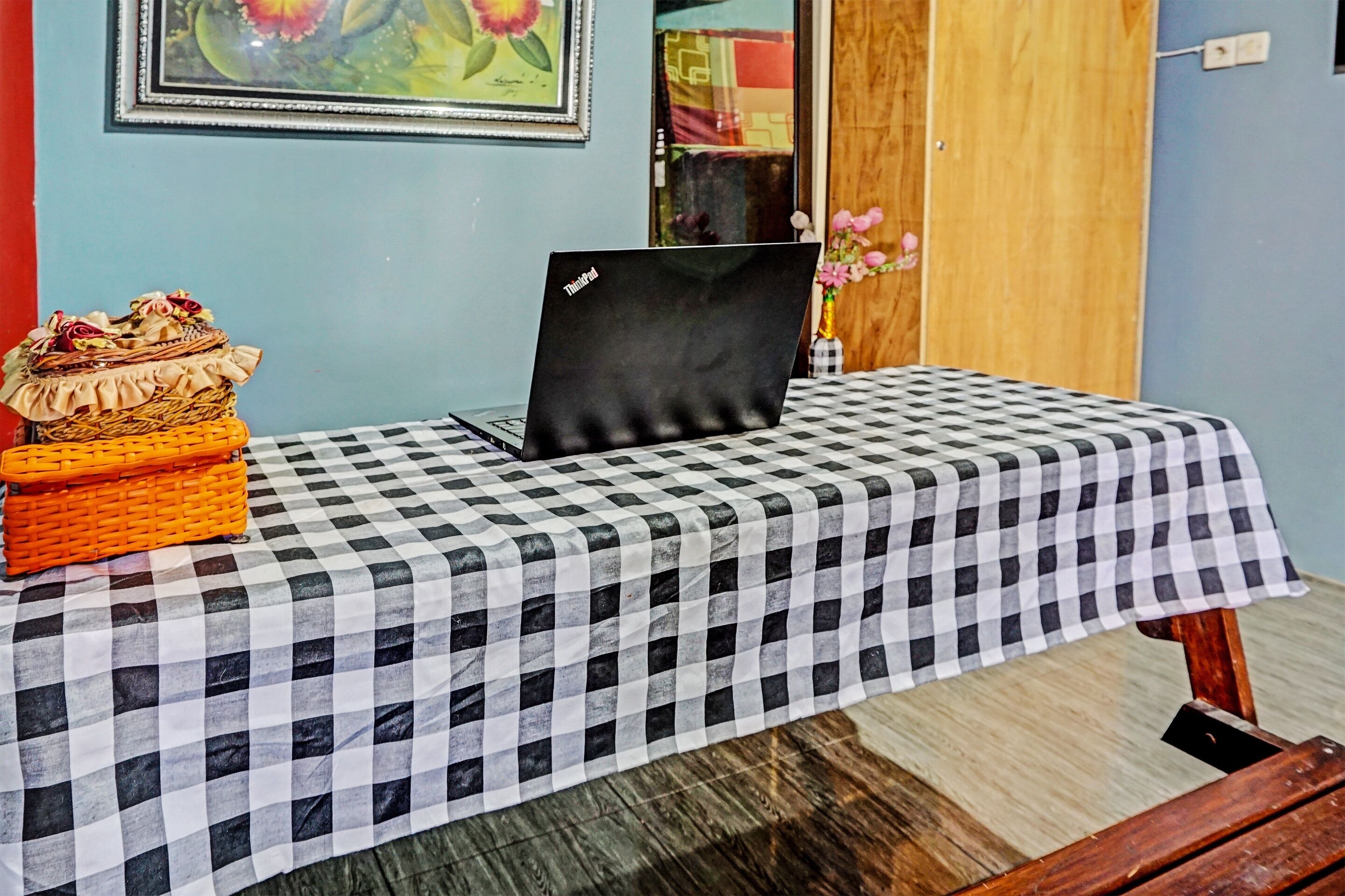 Ein Zimmer mit einem Tisch, der mit einer karierten Tischdecke bedeckt ist, einem Laptop, Korbwannen und einem gerahmten Blumenbild an der Wand.