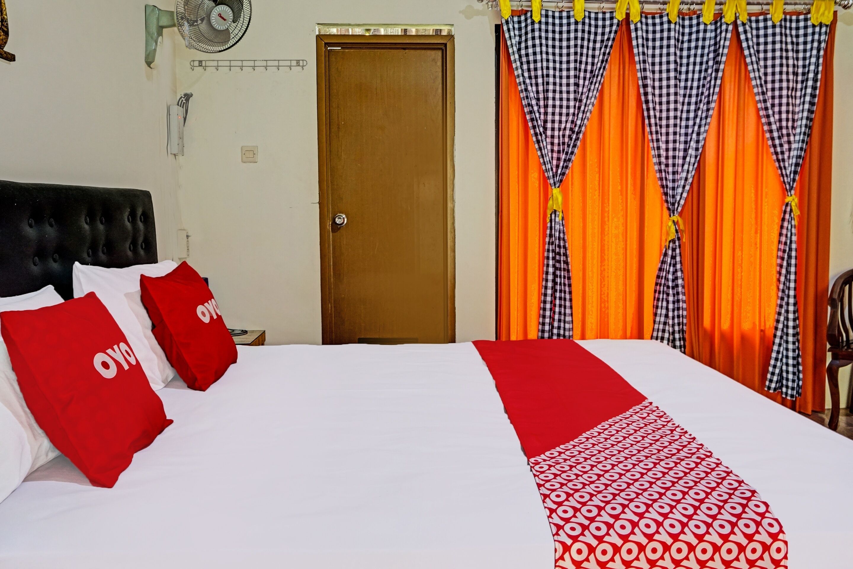 Une chambre d’hôtel avec un lit, une porte en bois, un ventilateur et des rideaux.