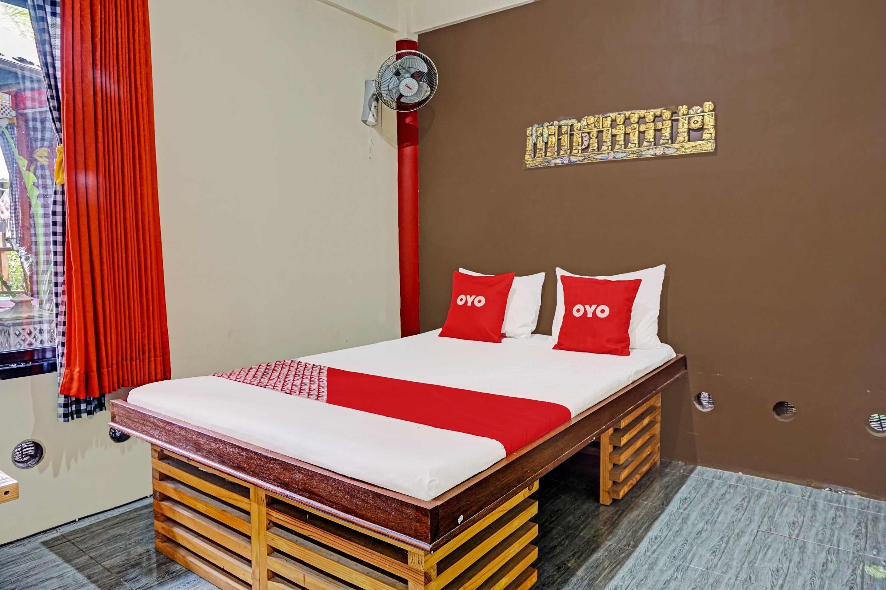 Une chambre d’hôtel avec un lit, une literie rouge et blanche, un ventilateur fixé au mur et un élément décoratif mural.
