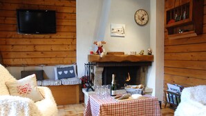 A cozy living room with a fireplace, a television mounted on the wall, and a checkered tablecloth-covered table with glasses and a bottle. - Appartement Avec vue Panoramique sur la Vallée (Le Grand-Bornand)