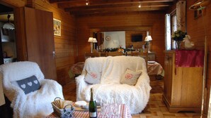A cozy wooden cabin interior with a plush white sofa, a dining table with a bottle and glasses, and a wooden cabinet. - Appartement Avec vue Panoramique sur la Vallée (Le Grand-Bornand)