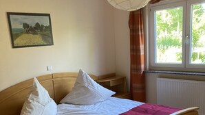Ett sovrum med en säng i trä, vita sängkläder, en röd pläd, ett nattduksbord, en lampa, ett fönster med gardiner och en tavla på väggen.