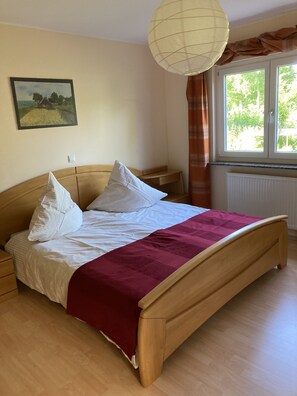 A bedroom with a wooden bed, white bedding, a red throw blanket, a nightstand, a lamp, a window with curtains, and a painting on the wall.