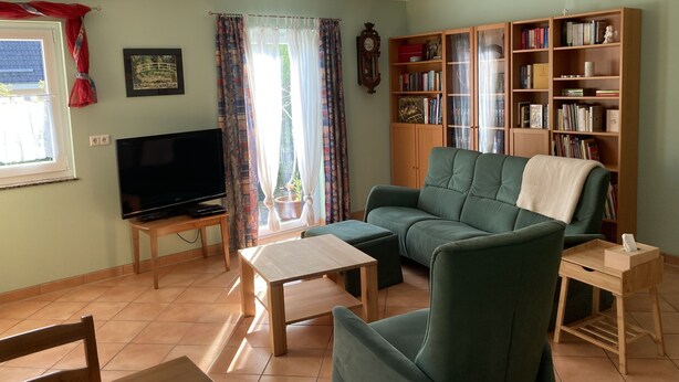 Ett vardagsrum med en grön soffa, ett soffbord i trä, en tv på ett tv-bord, en bokhylla och ett fönster med gardiner.