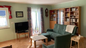 Una sala de estar con un sofá verde, una mesa de centro de madera, un televisor sobre un mueble bajo, una estantería y una ventana con cortinas.