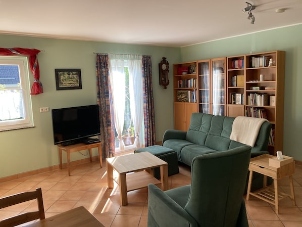 A living room with a green sofa, a wooden coffee table, a TV on a stand, a bookshelf, and a window with curtains.