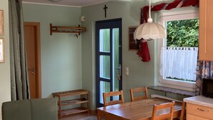En matplats med ett träbord och stolar, ett fönster med utsikt över grönområden, en dörr med en blå ram och en takfläkt.