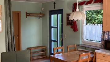 Un comedor con mesa y sillas de madera, ventana con vista a follaje, puerta con marco azul y ventilador de techo.