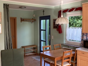 A dining area with a wooden table and chairs, a window with a view of greenery, a door with a blue frame, and a ceiling fan.