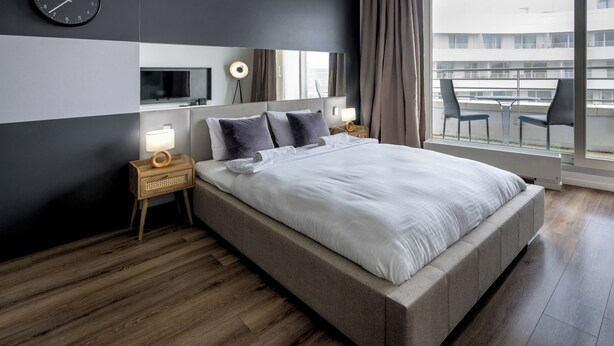Ein modernes Hotelzimmer mit einem großen Bett, einem Nachttisch mit Lampe, einer Wanduhr und einem großen Fenster mit Blick auf die Stadt.
