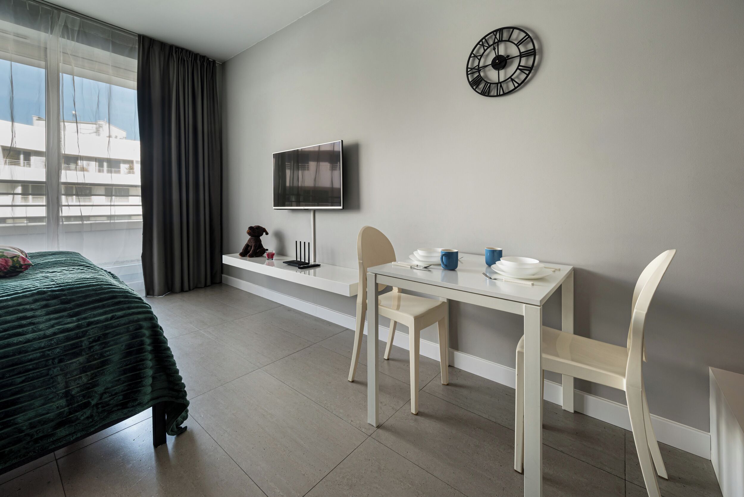 A hotel room with a bed, a desk with chairs, a flat-screen TV, a clock, and a window with curtains.