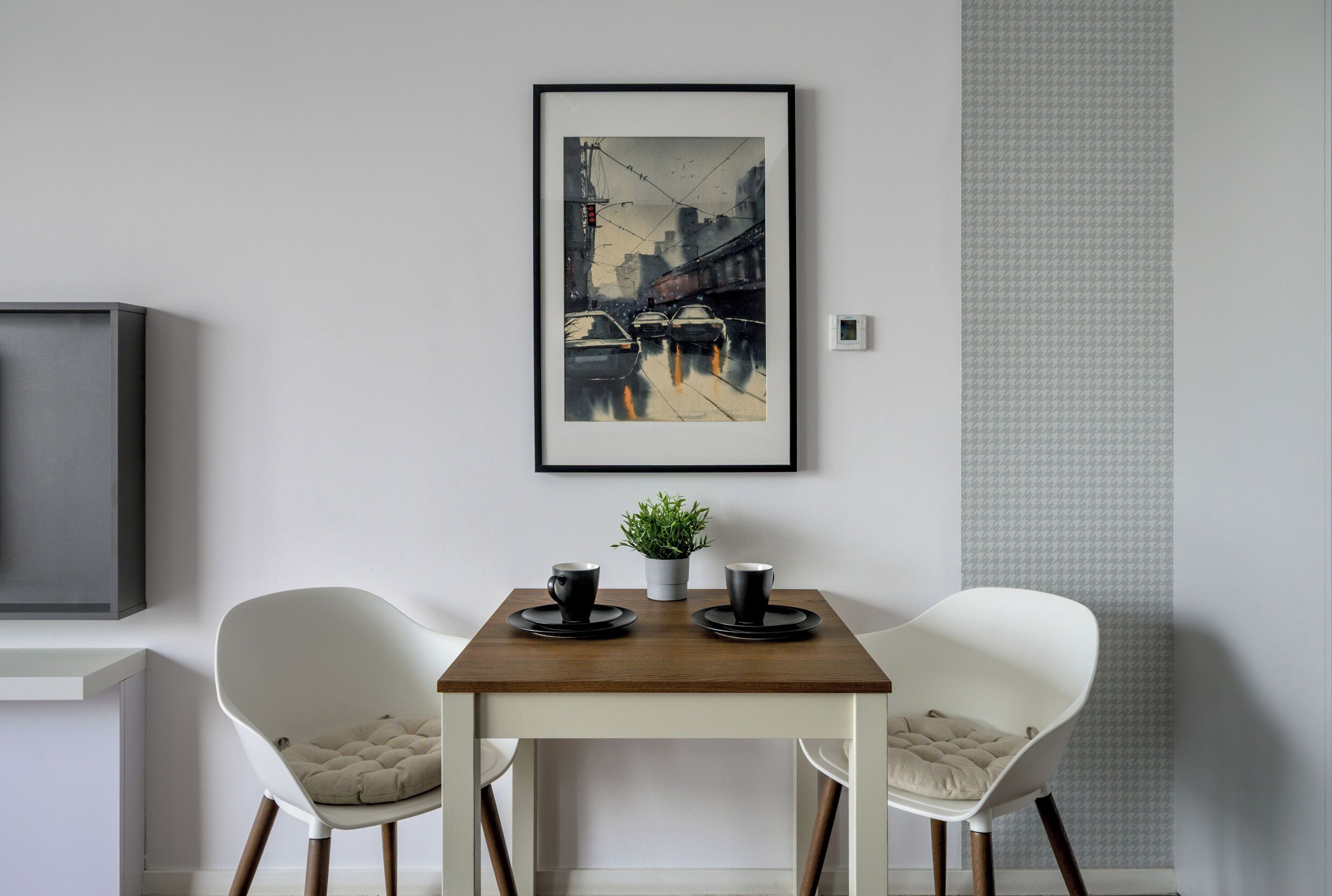 A modern dining area with a wooden table, two white chairs, and a framed cityscape painting above.