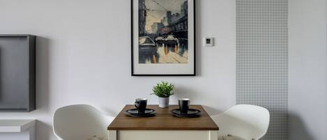 A modern dining area with a wooden table, two white chairs, and a framed cityscape painting above.