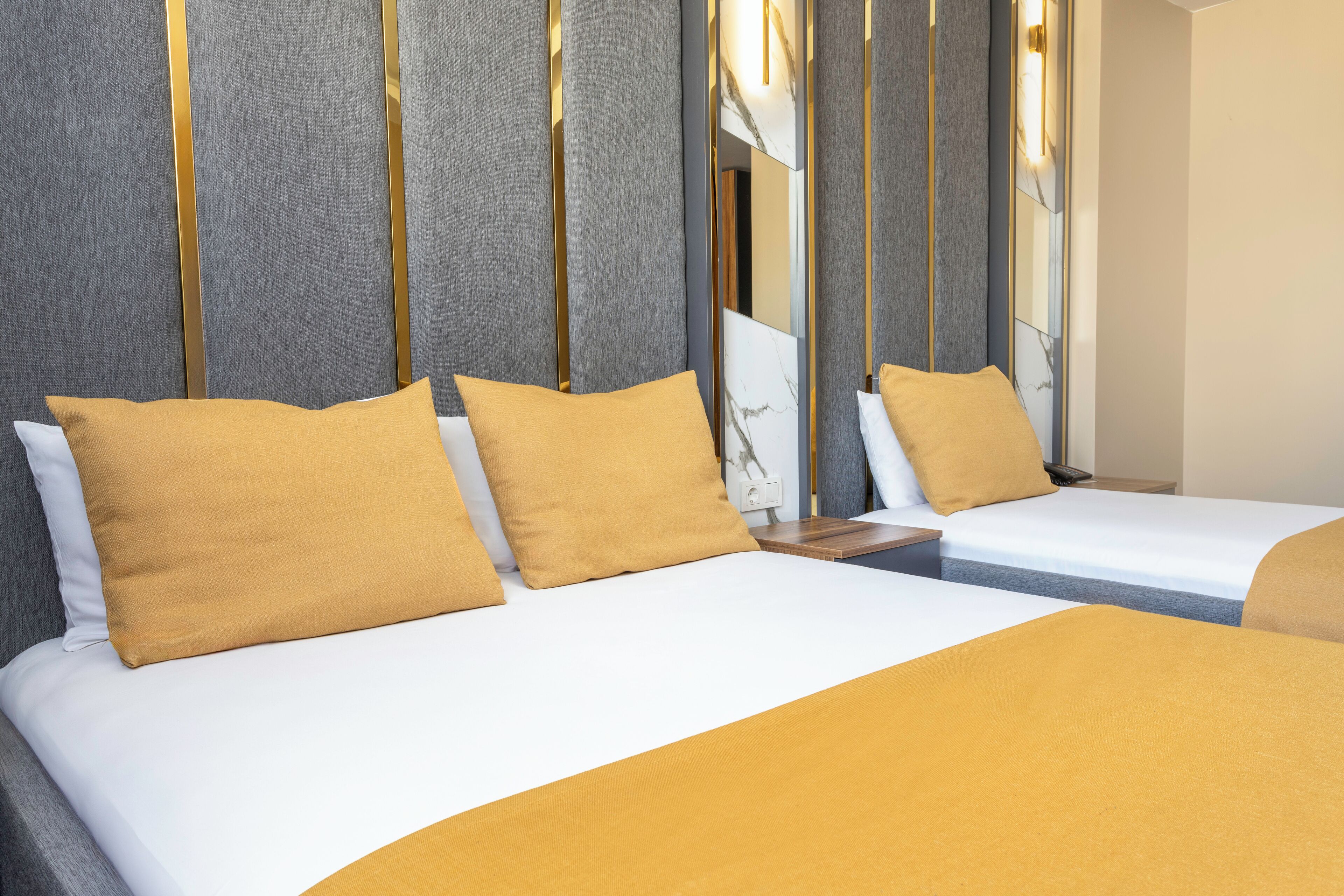 Habitación de hotel con dos camas, cada una con colchón blanco y manta color mostaza. La habitación presenta un diseño moderno con detalles verticales dorados en la pared y un espejo con estampado de mármol sobre las camas.