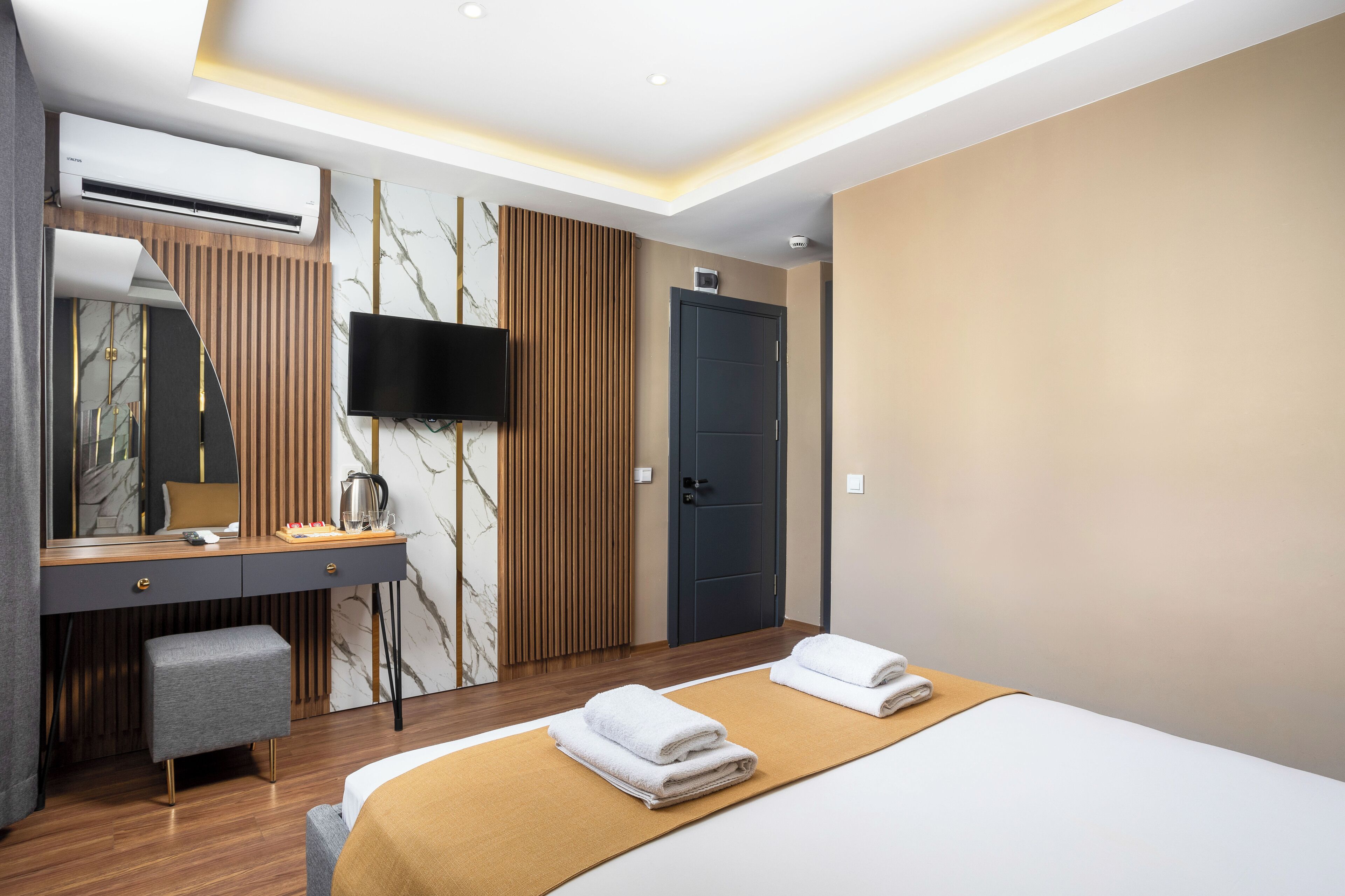 A modern hotel room with a bed, a desk with a chair, a flat-screen TV, an air conditioner, and a door.