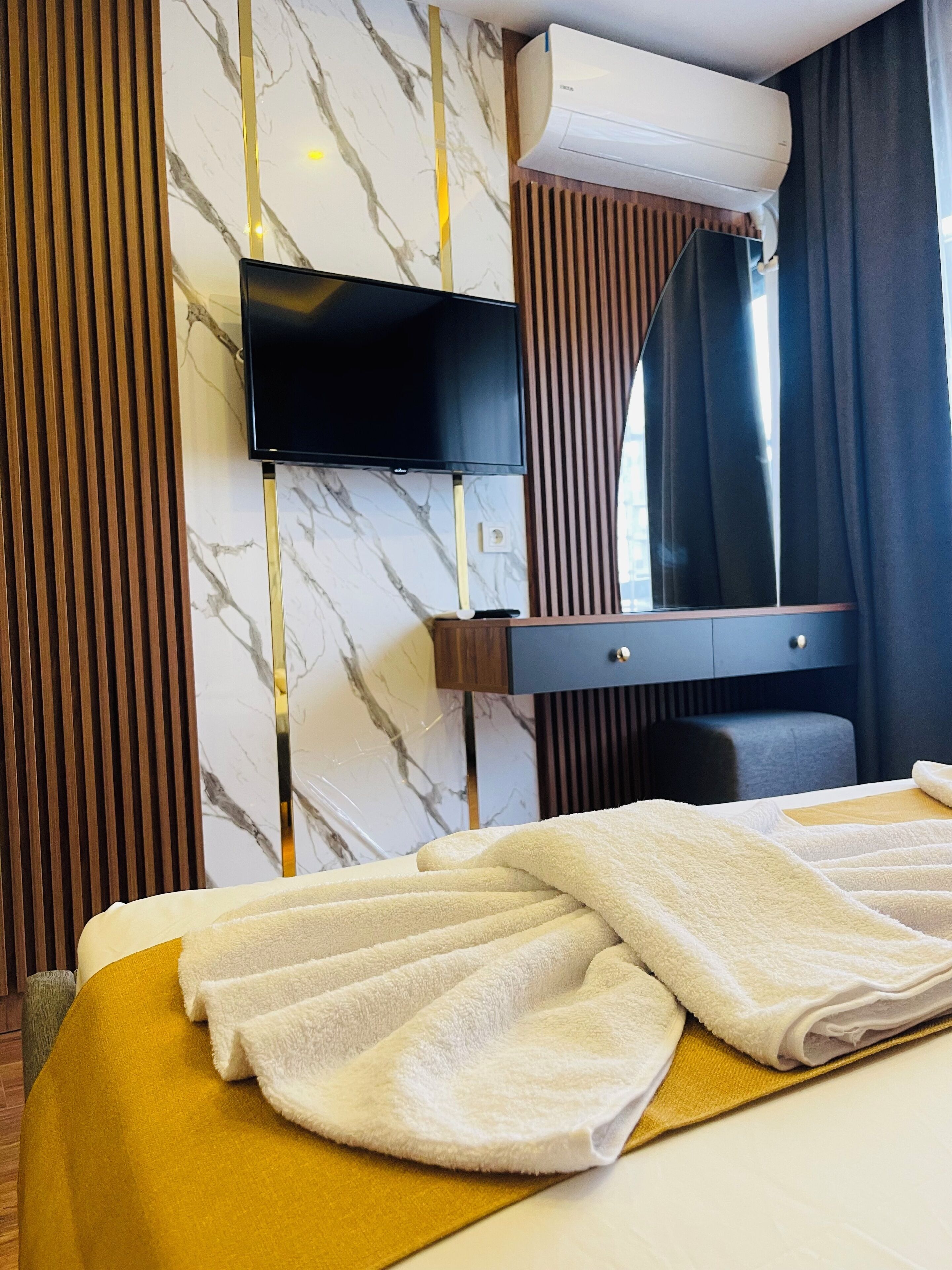 A hotel room with a bed, a flat-screen TV mounted on the wall, a mirror, and a window with curtains.