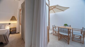 A bedroom with a bed, bedside table, lamp, and curtains. There is a dining area with a table, chairs, and a plant. - Hotel Tres Tocs (Ciutadella de Menorca)