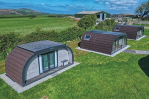 A row of modern, semi-open structures with glass walls, set in a lush green field with a distant view of hills and a clear sky.