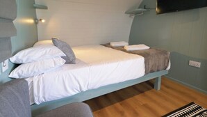 A single bed with white bedding and a grey pillow, a small bedside table, a mounted TV, and a power outlet on the wall. - Ocean View 3 - 1 Bed Pod - Llanrhidian (Swansea)