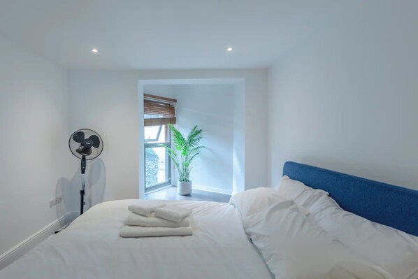 A modern bedroom with a neatly made bed, a standing fan, a window with blinds, and a potted plant.