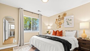 A bedroom with a large bed, a nightstand, a lamp, a mirror, and a window with curtains.