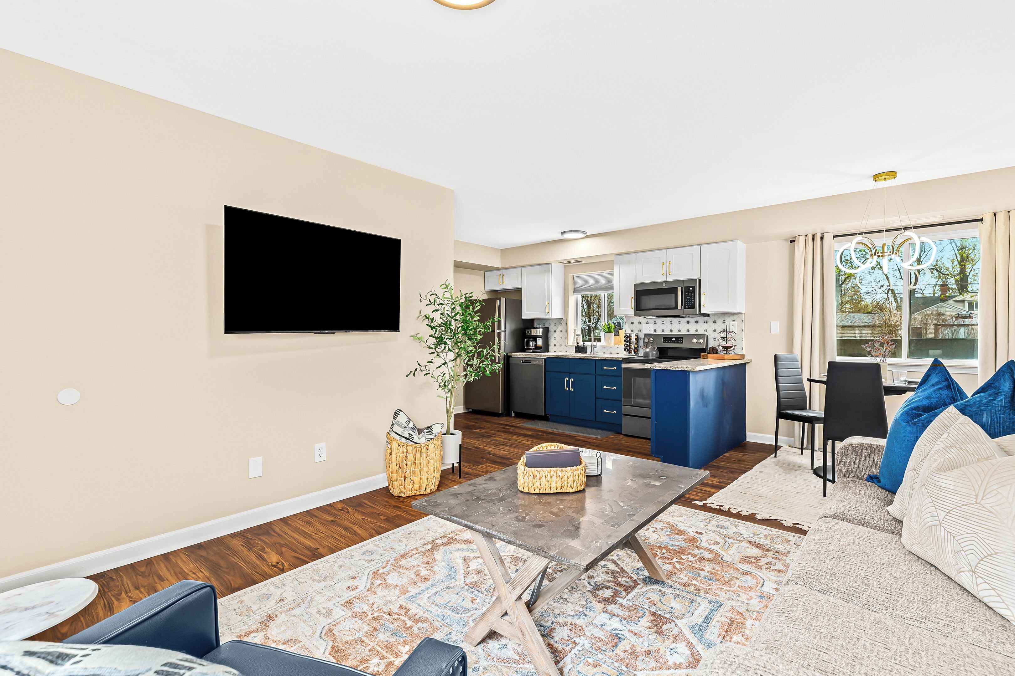 A modern living room with a flat-screen TV, a coffee table, and a sofa. There is a kitchen in the background with blue cabinets and a dining area with a table and chairs.