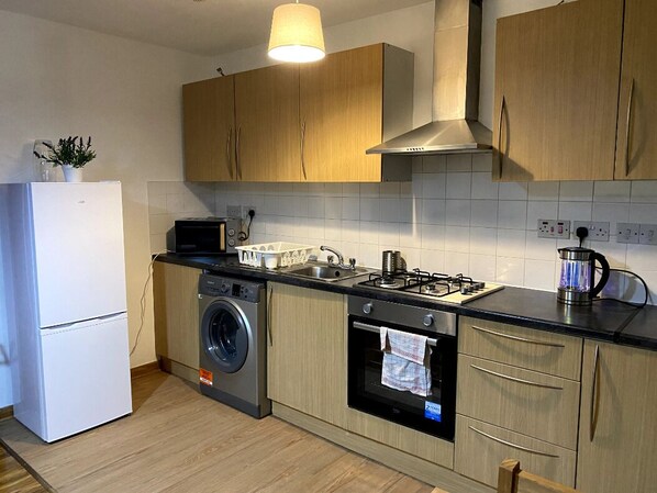 Compact kitchen with washing machine, oven, and gas stove, featuring light wood cabinetry and a black countertop.