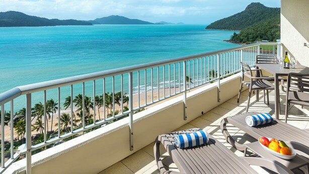 A balcony with a view of a beach and ocean, furnished with a table, chairs, and a fruit bowl. - Whitsunday Apartment West 1302 (Hamilton Island)