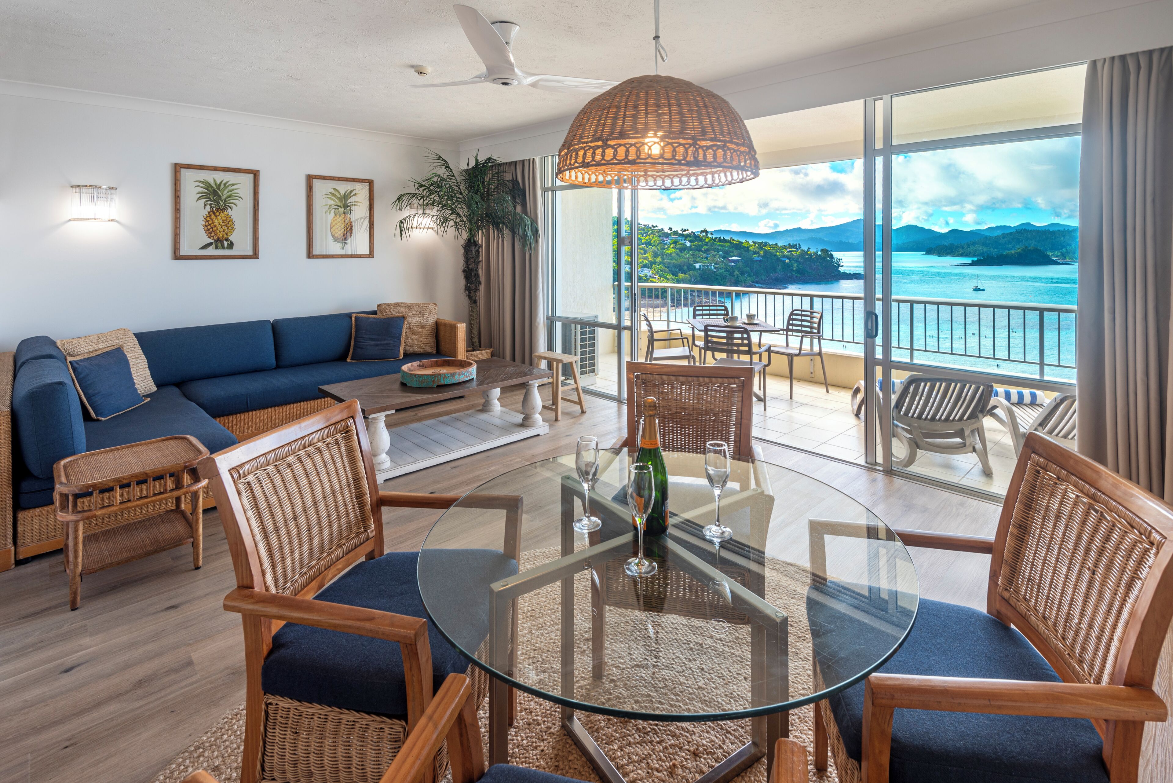 Een moderne woonkamer met een glazen salontafel, rieten stoelen, een bank en een balkon met uitzicht op zee.