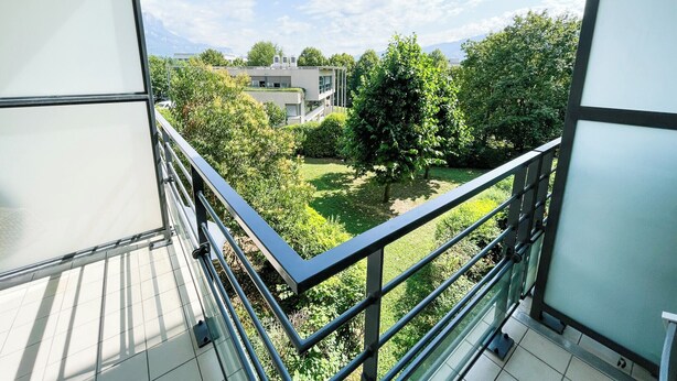 A balcony with a metal railing overlooking a grassy area with trees and a building in the background.